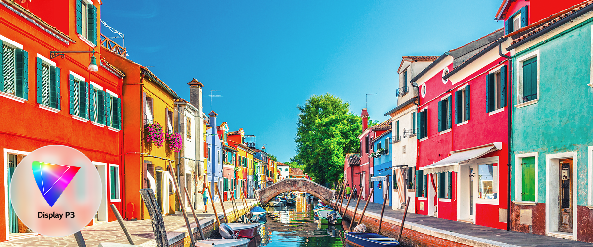 A side-by-side comparison of 'Conventional IPS' on the left and 'Neo:LED' on the right. Both screens display an image of Burano Island, Italy, with the Neo:LED display showing more vivid and rich colors. The conventional IPS features an icon in the lower-left corner indicating support for the Adobe RGB color gamut, whereas the Neo:LED has an icon in the lower-right corner that signifies support for both Adobe RGB and Display P3 color gamuts.
