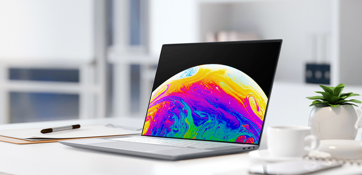 A laptop placed on a modern and minimalist office desk highlights its high-resolution display. The screen features a vibrant, abstract cosmic image, emphasizing its vivid color reproduction. The desk is decorated with a small plant, a notebook, and a pen, evoking a productive workspace.